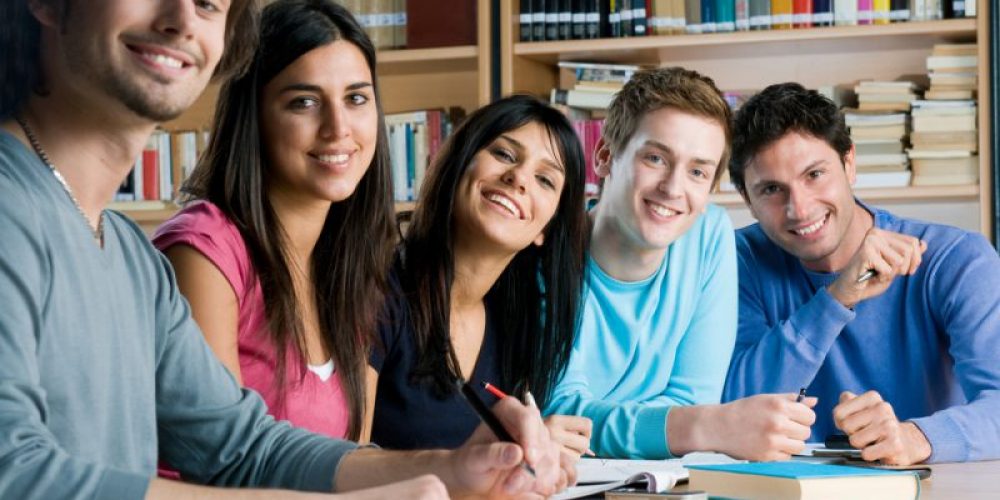 Happy group of young students studying together in a college library and looking at camera smiling