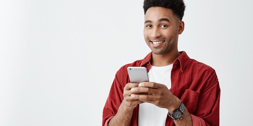 Positive emotions. Close up of young good-looking dark-skinned male with afro hairstyle in white t-shirt and red shirt smiling with teeth, chatting with friend on smartphone.